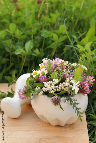 Ceramic mortar with pestle, different wildflowers and herbs on wooden board in meadow