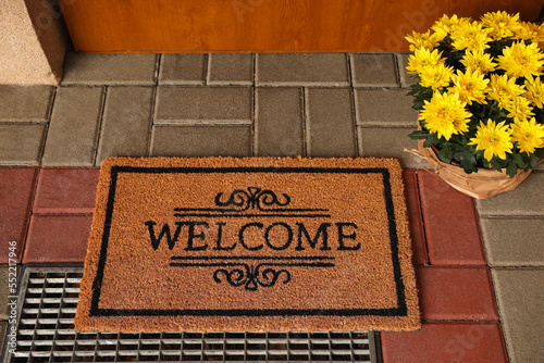 Door mat with word Welcome and beautiful flowers near entrance