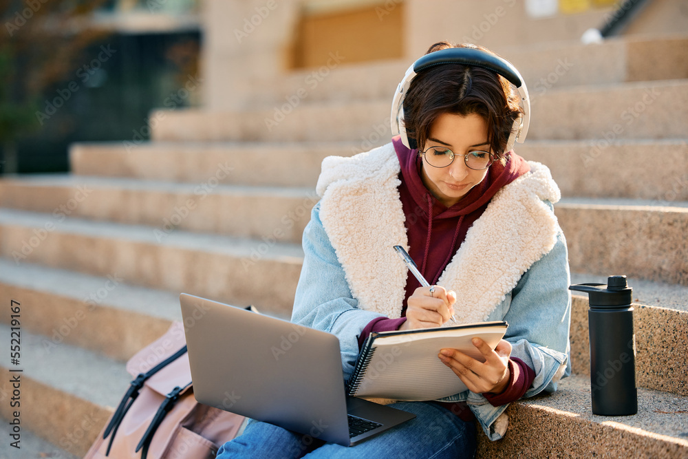 Young student using laptop and writing in her notebook while learning at campus.