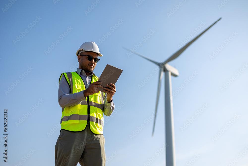 Engineer India man working with tablet at windmill farm Generating electricity clean energy. Wind turbine farm generator by alternative green energy. Asian engineer checking control electric power Engineer India man working with tablet at windmill farm Generating electricity clean energy. Wind turbine farm generator by alternative green energy. Asian engineer checking control electric power