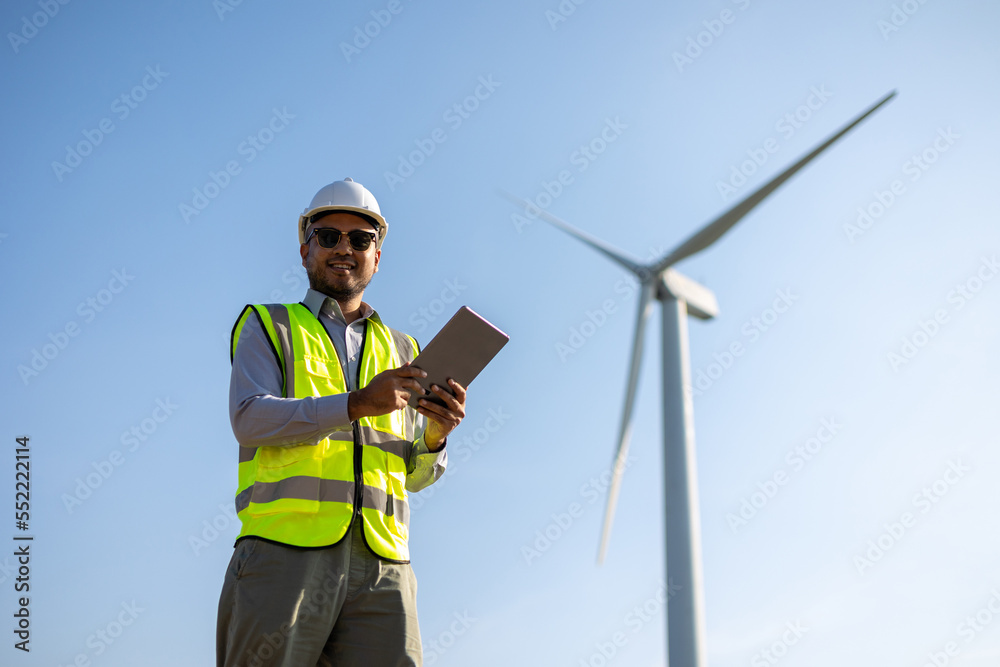 Engineer India man working with tablet at windmill farm Generating electricity clean energy. Wind turbine farm generator by alternative green energy. Asian engineer checking control electric power Engineer India man working with tablet at windmill farm Generating electricity clean energy. Wind turbine farm generator by alternative green energy. Asian engineer checking control electric power