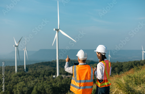 Two Engineer man and woman working at windmill farm Generating electricity clean energy. Wind turbine farm generator by alternative green energy. Asian engineer checking control electric power