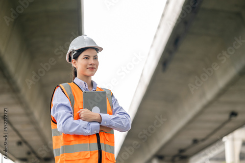 Asian engineer worker woman or architect looking construction with tablet wear white safety helmet in construction site. Standing at highway concrete road site. Progress planning of highway bridge.