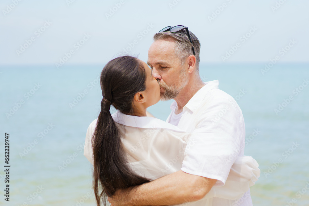 Happy Asian and caucasian white couple enjoy relaxing together at the beach during vacation or their honeymoon dating. Husband gently kissing his wife. Happy Asian and caucasian white couple enjoy relaxing together at the beach during vacation or their honeymoon dating. Husband gently kissing his wife.