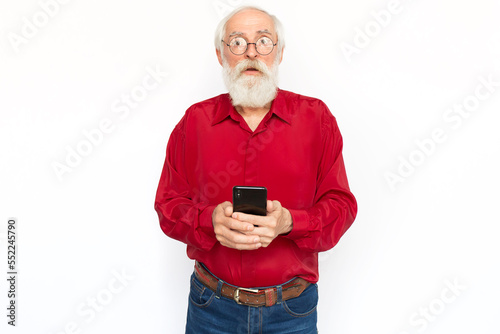 Focused senior man learning to use smartphone. Upset Caucasian male model with gray hair and beard in red shirt and glasses looking at camera, mouth open, asking for help. Modern technology concept