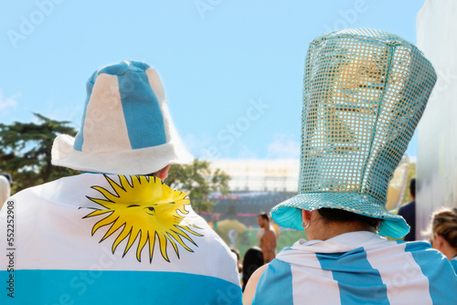 Man and woman fans of the Argentine national team with their backs to the camera watch the game on TV.