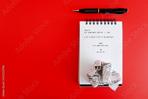 Pen , crumpled paper and  notebook with screenwriting scene written, on red copy space background. Concept of  professional screenwriter job, TV or movie film script writing or writing block