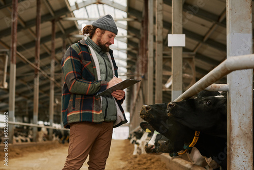 Farmer standing in front of cows in big modern barn making notes in document