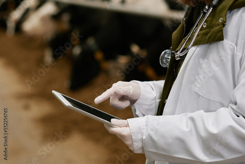 Close-up of veterinarian in white coat typing on digital tablet while working with animals on farm