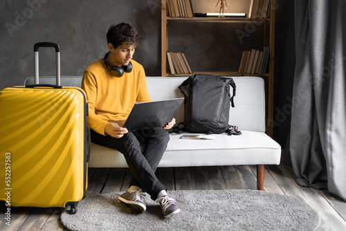 Young man traveler planning vacation trip and using computer to book hotel room online. Male tourist wearing sweater sitting on sofa and browsing rental holiday accommodation website