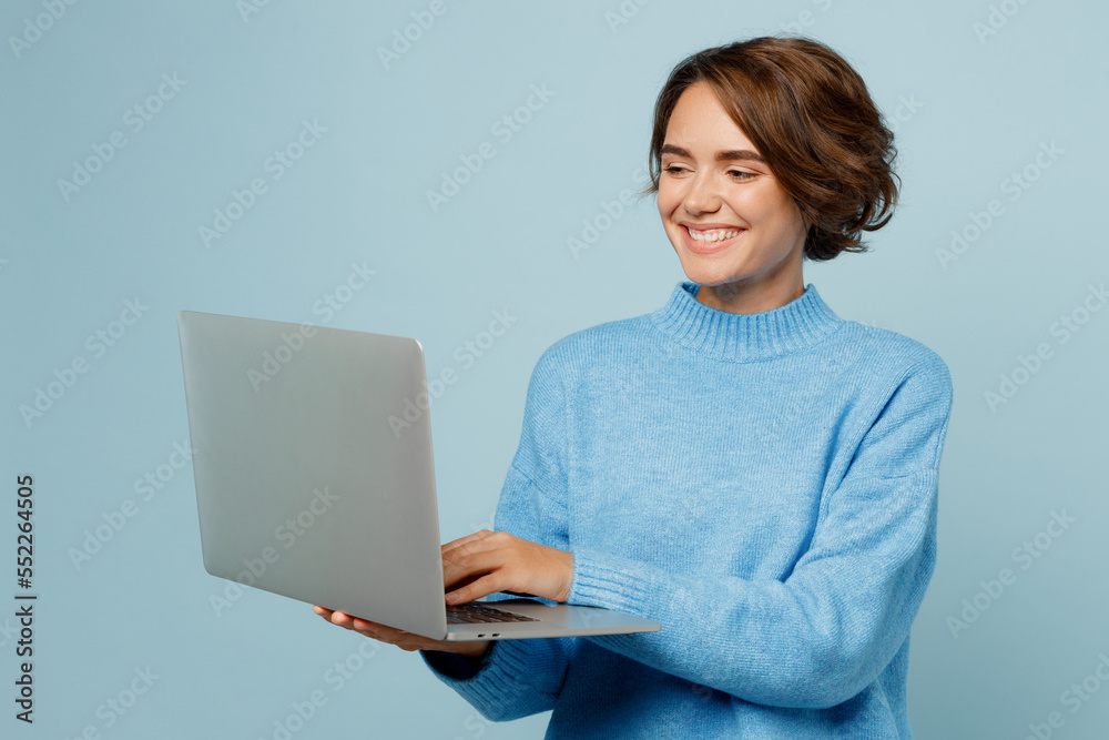Young smiling happy fun caucasian smart IT woman wear knitted sweater hold use work on laptop pc computer isolated on plain pastel light blue cyan background studio portrait. People lifestyle concept. Young smiling happy fun caucasian smart IT woman wear knitted sweater hold use work on laptop pc computer isolated on plain pastel light blue cyan background studio portrait. People lifestyle concept.