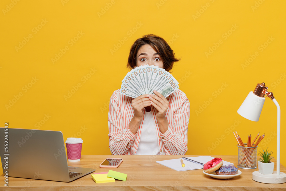 Young employee business woman wears casual shirt sit work at office desk cover mouth with fan of cash money in dollar banknotes isolated on plain yellow color background. Achievement career concept. Young employee business woman wears casual shirt sit work at office desk cover mouth with fan of cash money in dollar banknotes isolated on plain yellow color background. Achievement career concept.