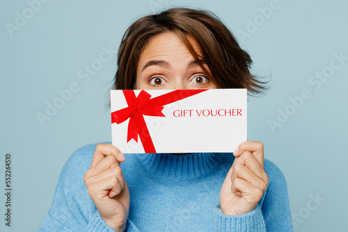 Close up young happy surprised shocked woman wear knitted sweater holding covering mouth with gift certificate coupon voucher card for store isolated on plain pastel light blue cyan background studio.