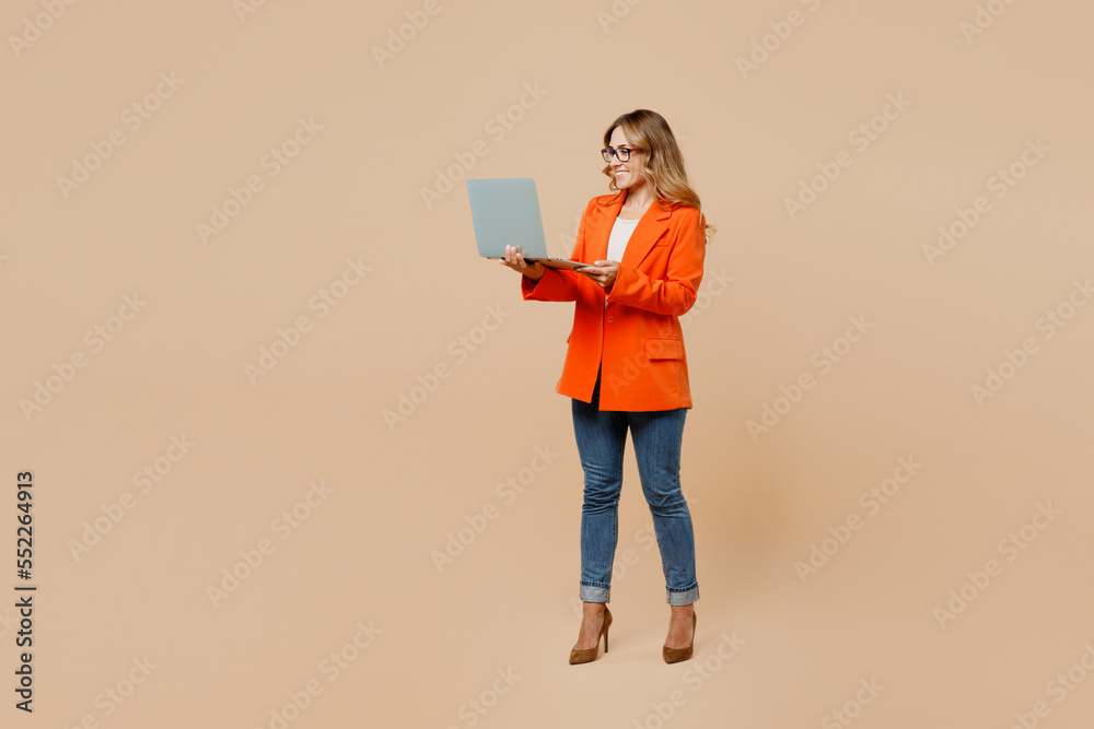 Full body confident young successful employee IT business woman corporate lawyer wear classic formal orange suit glasses work in office hold use laptop pc computer isolated on plain beige background. Full body confident young successful employee IT business woman corporate lawyer wear classic formal orange suit glasses work in office hold use laptop pc computer isolated on plain beige background.