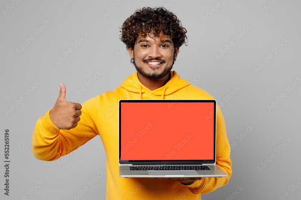 Young smiling happy fun IT Indian man 20s wear casual yellow hoody hold use work on laptop pc computer with blank screen workspace area show thumb up isolated on plain grey background studio portrait. Young smiling happy fun IT Indian man 20s wear casual yellow hoody hold use work on laptop pc computer with blank screen workspace area show thumb up isolated on plain grey background studio portrait.