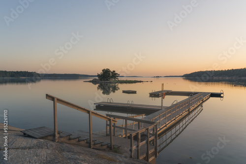 Nagu, Finland - 12.06.2022: the quiet shore of the bay after sunset. A line of trees and a small island on the horizon. A rocky shore with a pier and a bench in the foreground. Nature of Finland.