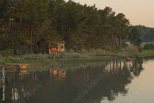 Nagu, Finland - 12.06.2022: A small red house on the shore of the evening bay. The pine tree line is reflected in the water. Nature of Finland. Scandinavia.