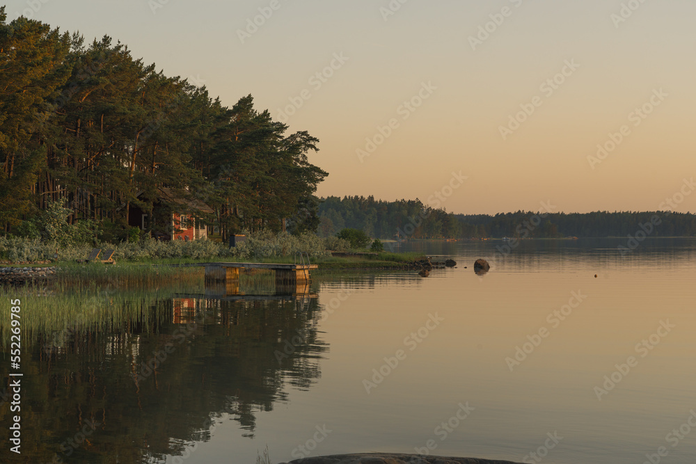 Nagu, Finland - 12.06.2022: A small red house on the shore of the evening bay. The pine tree line is reflected in the water. Nature of Finland. Scandinavia.