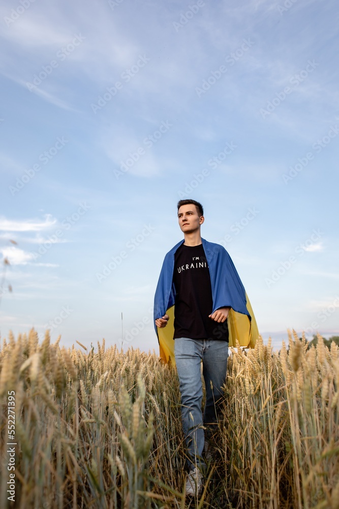 a young guy is walking in a wheat field against the background of a blue sky with textured clouds at sunset with the Ukrainian flag on his shoulders a young guy is walking in a wheat field against the background of a blue sky with textured clouds at sunset with the Ukrainian flag on his shoulders