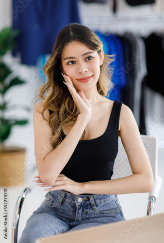 Millennial Asian young beautiful sexy female customer in black tank top and jeans outfit sitting on chair waiting for professional dressmaker designer seamstress in tailor studio workshop office