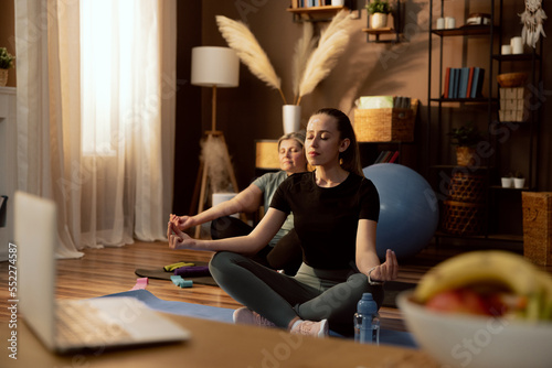 Young daughter with elderly mother in background sitting on yoga mat in otos pose closing eyes in front of laptop with yoga lesson online relaxing. photo