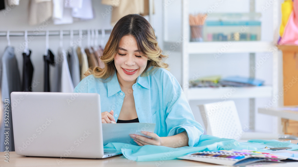Millennial Asian young beautiful professional female designer dressmaker sitting smiling at working table with clothing color pattern sample and fabric using laptop computer in tailor studio workshop Millennial Asian young beautiful professional female designer dressmaker sitting smiling at working table with clothing color pattern sample and fabric using laptop computer in tailor studio workshop