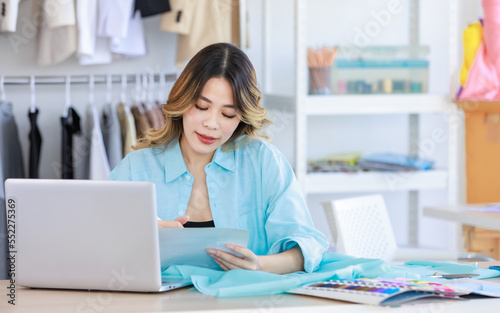 Millennial Asian young beautiful professional female designer dressmaker sitting smiling at working table with clothing color pattern sample and fabric using laptop computer in tailor studio workshop
