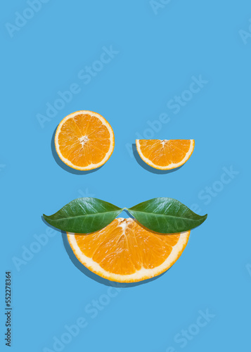 Creative summer face with eyes made of orange fruit slices and mustache made of leaves on bright blue vibrant background. Orange juice minimal tropic concept. Winking eye funny idea