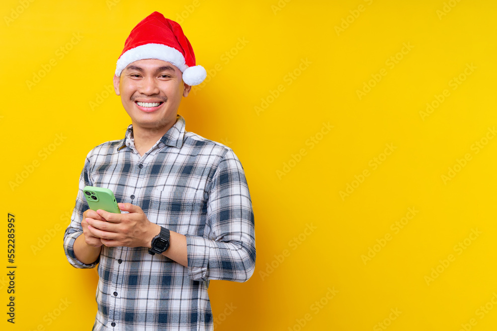 Smiling young Asian man in plaid shirt wearing Christmas hat holding mobile phone and looking confident at camera on yellow background. Christmas and Happy New Year 2023 holiday concept