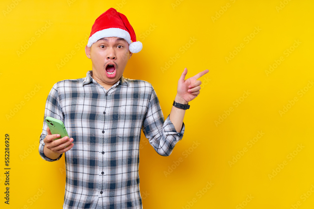 Surprised young Asian man in plaid shirt wearing Christmas hat holding mobile phone and pointing index finger aside isolated on yellow background. Christmas and Happy New Year 2023 holiday concept Surprised young Asian man in plaid shirt wearing Christmas hat holding mobile phone and pointing index finger aside isolated on yellow background. Christmas and Happy New Year 2023 holiday concept