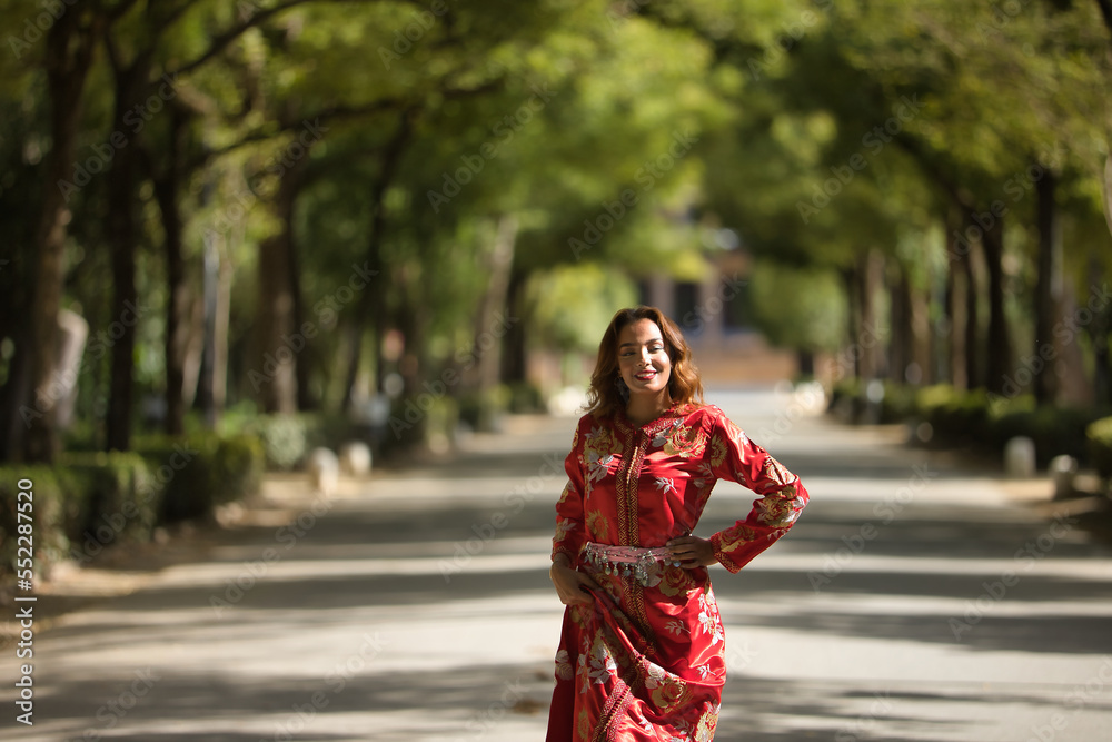 Beautiful young woman in a typical Moroccan red suit, embroidered with gold and silver threads, posing in an outdoor park avenue. Concept beauty, ethnicity, typical suits, Marrakech, Arab. Beautiful young woman in a typical Moroccan red suit, embroidered with gold and silver threads, posing in an outdoor park avenue. Concept beauty, ethnicity, typical suits, Marrakech, Arab.