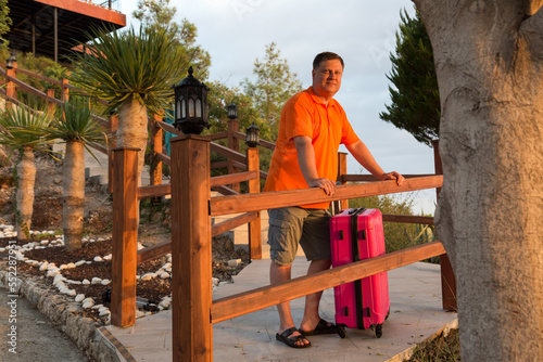 An adult tourist in shorts with a large suitcase looks at the sunset.