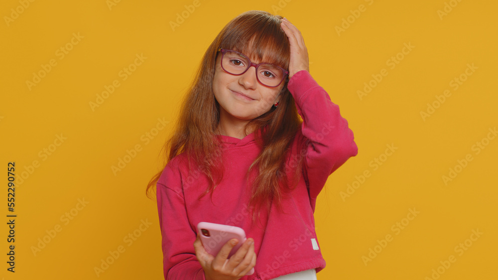 Young toddler school girl in glasses use smartphone typing browsing, loses becoming surprised sudden lottery results, bad fortune, loss. Education, study. Teen female child kid on yellow background Young toddler school girl in glasses use smartphone typing browsing, loses becoming surprised sudden lottery results, bad fortune, loss. Education, study. Teen female child kid on yellow background
