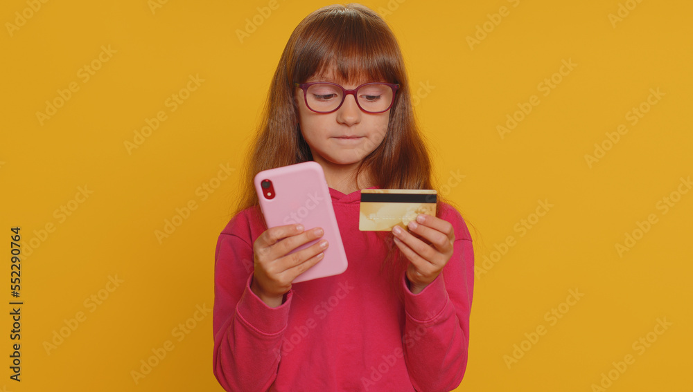 Young preteen child girl kid using credit bank card and smartphone while transferring money, purchases online shopping, order food delivery. Little toddler children alone on studio yellow background Young preteen child girl kid using credit bank card and smartphone while transferring money, purchases online shopping, order food delivery. Little toddler children alone on studio yellow background