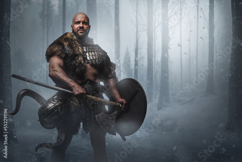 Studio shot of handsome ancient viking holding axe and shield in forest in snowfall.