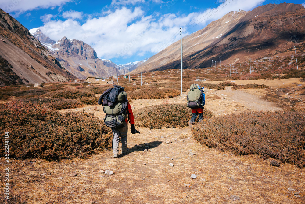 Two Sherpas from behind carry their luggage through the Annapurna Mountains. Two trekkers hike through the barren landscape of the Himalayas in Nepal