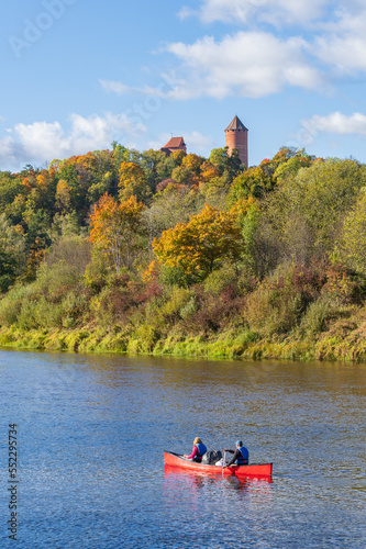 Red canoe swimming in river of Gauja on sunny autumn day. Turaida Castle and forest in autumn colorful foliage on river bank