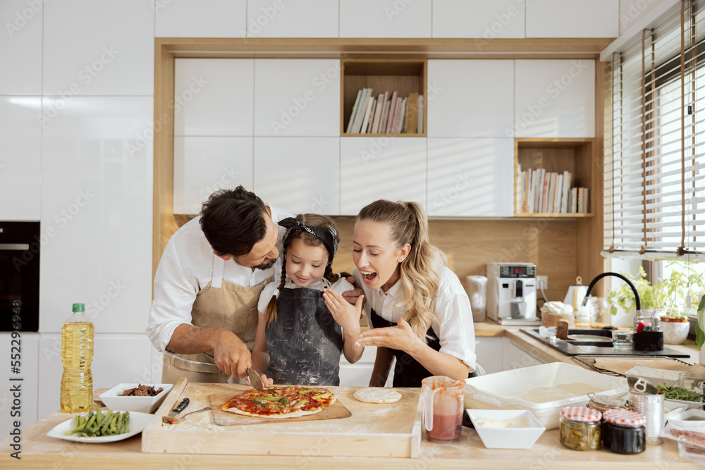 Delighted kid child daughter cutting homemade pizza in pieces wearing aprons. happy parents family spending time together in modern kitchen with large window. Delighted kid child daughter cutting homemade pizza in pieces wearing aprons. happy parents family spending time together in modern kitchen with large window.