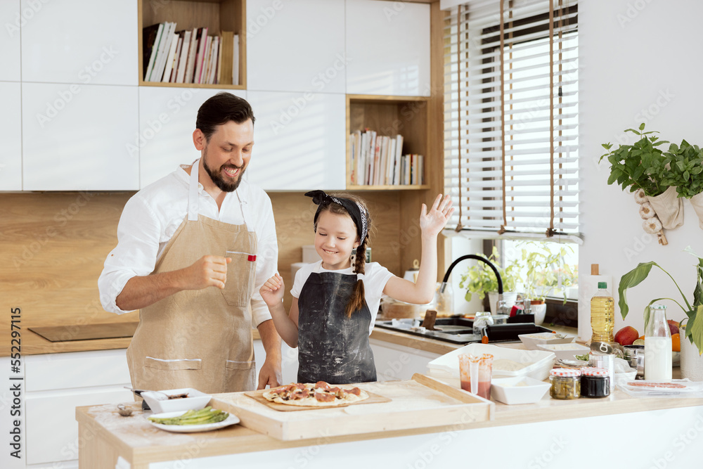 Delighted father and curious daughter laughing smiling wearing aprons looking at homemade baked pizza. Waiting for tasting having family dinner in modern kitchen. Delighted father and curious daughter laughing smiling wearing aprons looking at homemade baked pizza. Waiting for tasting having family dinner in modern kitchen.