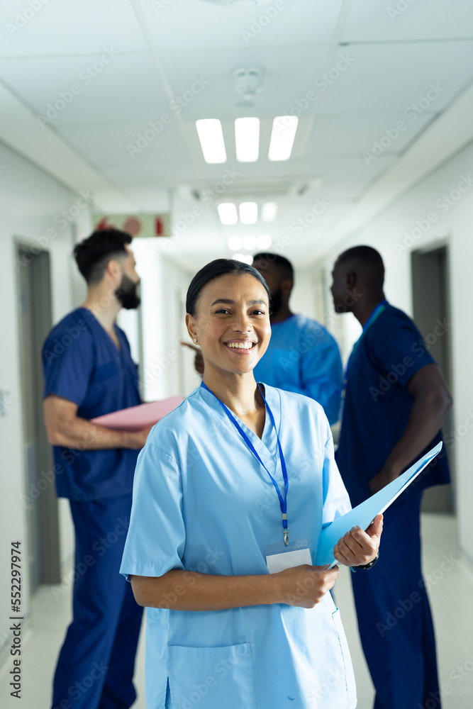 Fototapeta premium Vertical portrait of smiling biracial female healthcare worker in busy hospital corridor, copy space