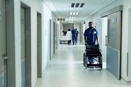Biracial male healthcare worker pushing empty wheelchair in busy hospital corridor
