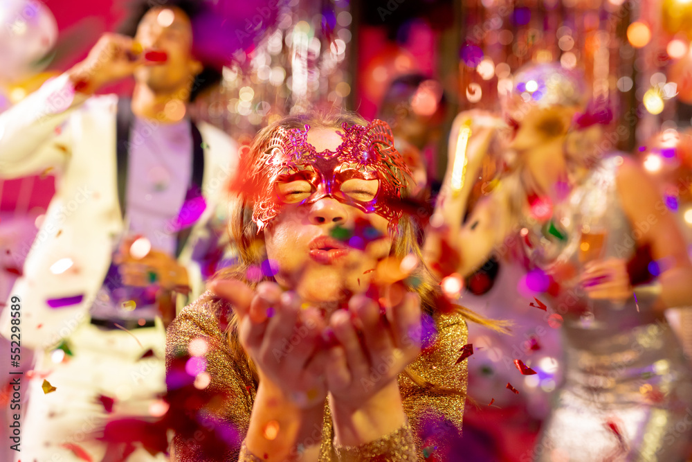 Happy caucasian woman in mask blowing glitter to camera at a party in a nightclub