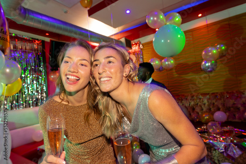 Two happy caucasian female friends embracing and drinking glasses of champagne at a nightclub