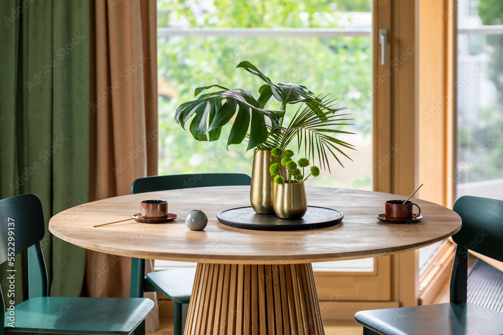 Fototapeta premium Creative composition of dinning room interior with wooden round table, gold vase with dried flowers, black trace, gray chairs, curtain, cups, window and personal accessories. Home decor. Template.