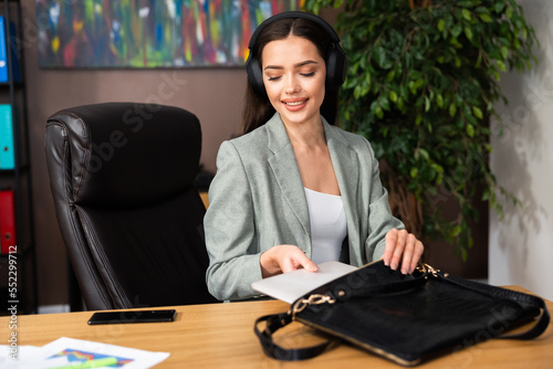 Delighted excited young woman in headphones smiling while putting laptop computer in bag after busy dayat work in cozy modern office. photo