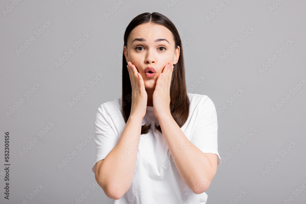 Portrait of young woman girl wearing white t shirt standing on grey background showing surprised emotions holding hands near face opening mouth. Portrait of young woman girl wearing white t shirt standing on grey background showing surprised emotions holding hands near face opening mouth.