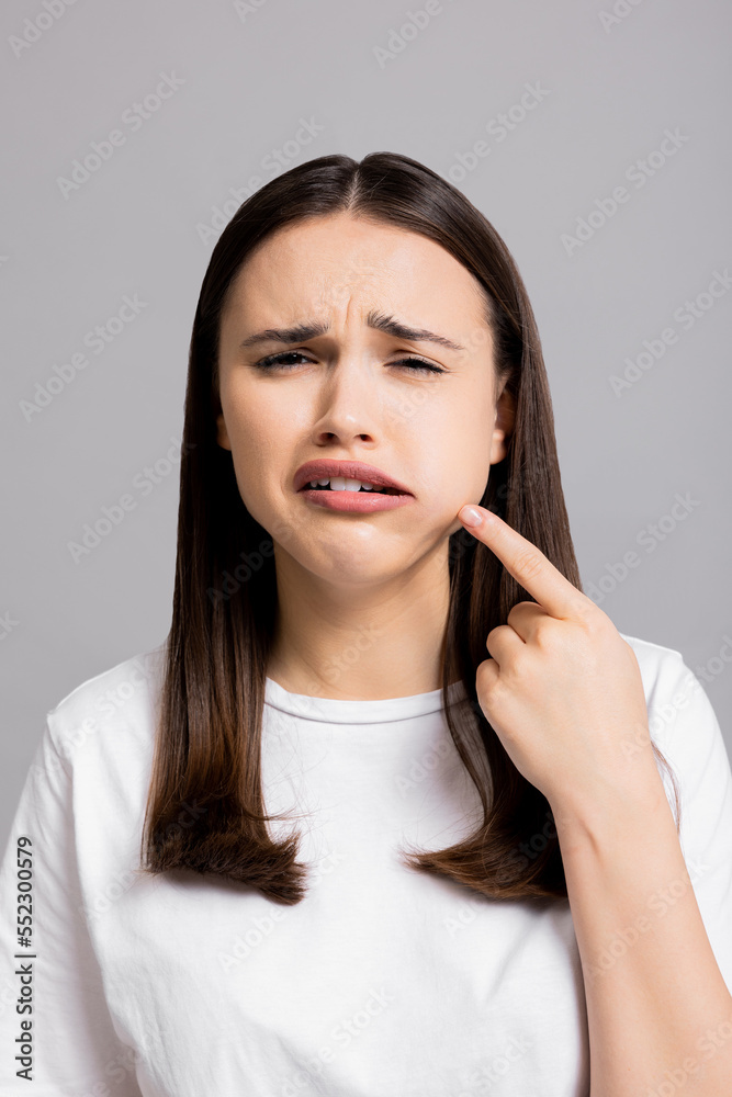 Crying tired sad woman suffers from painful toothache dental illness pointing on teeth standing on grey background in studio isolated wearing basic white t-shirt. Crying tired sad woman suffers from painful toothache dental illness pointing on teeth standing on grey background in studio isolated wearing basic white t-shirt.
