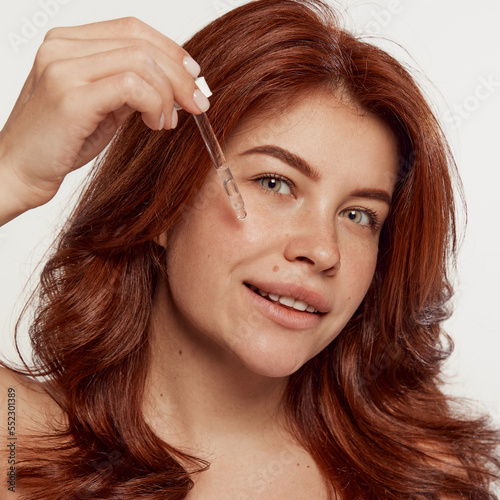 Portrait of young, beautiful, red-haired woman taking care after skin, applying face serum isolated on white background