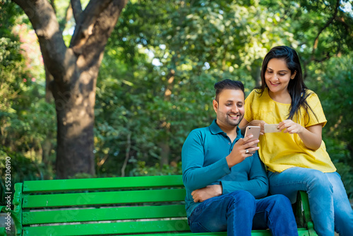 Indian man and woman using smartphone and bank card at park.