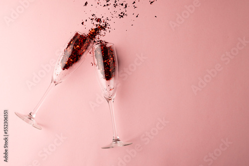 Two champagne glasses spilling red glitter confetti on pale pink background with copy space
